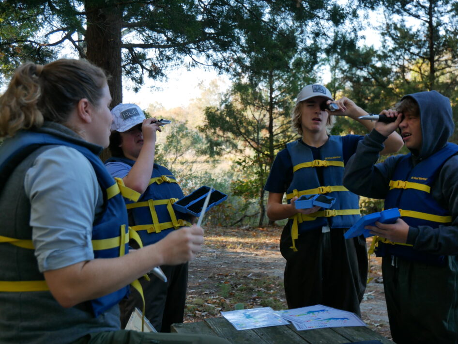 Four students wearing lifejackets hold refractometers while standing outdoors.