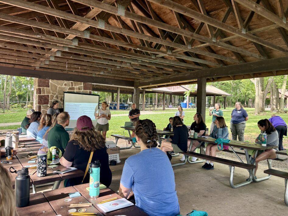 Educators sit at picnic tables under an outdoor pavilion, facing two standing presenters and a projection screen with a presentation.