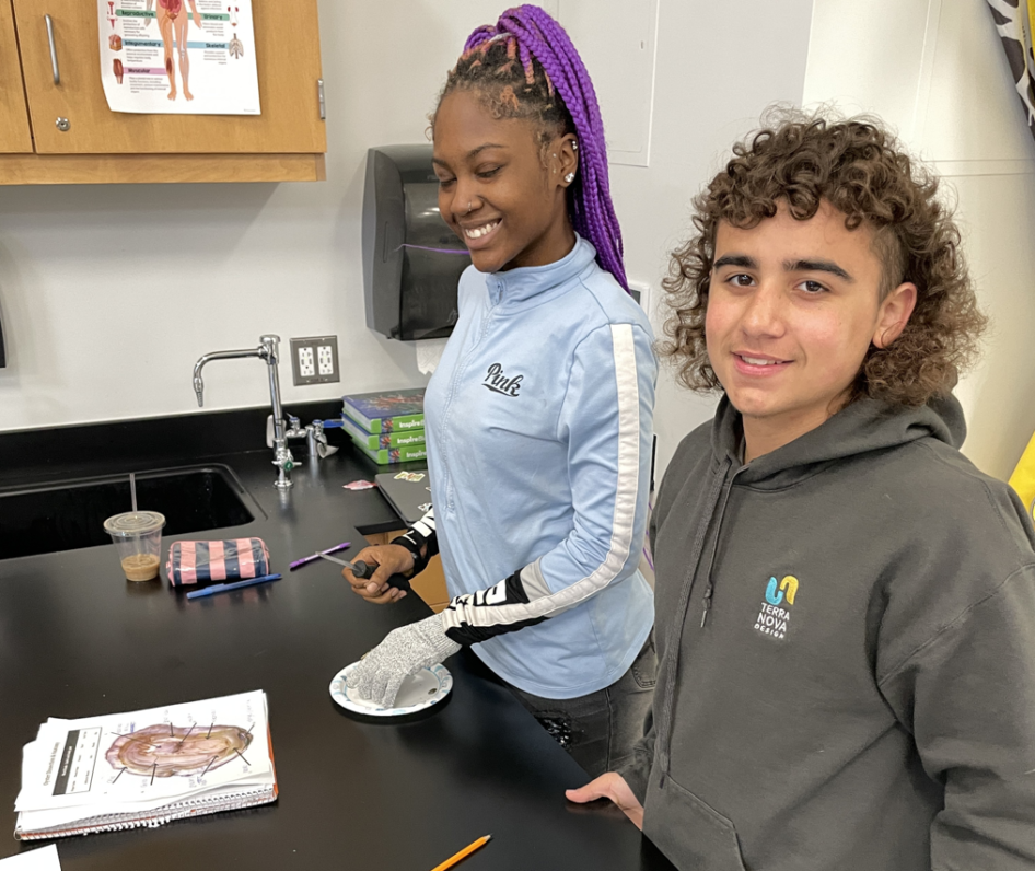 Two students stand in front of a black desk with a worksheet depicting the anatomy of an oyster.