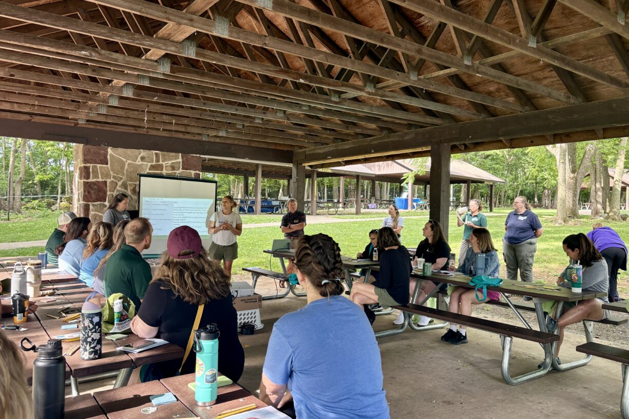 Educators sit at picnic tables under an outdoor pavilion, facing two standing presenters and a projection screen with a presentation.